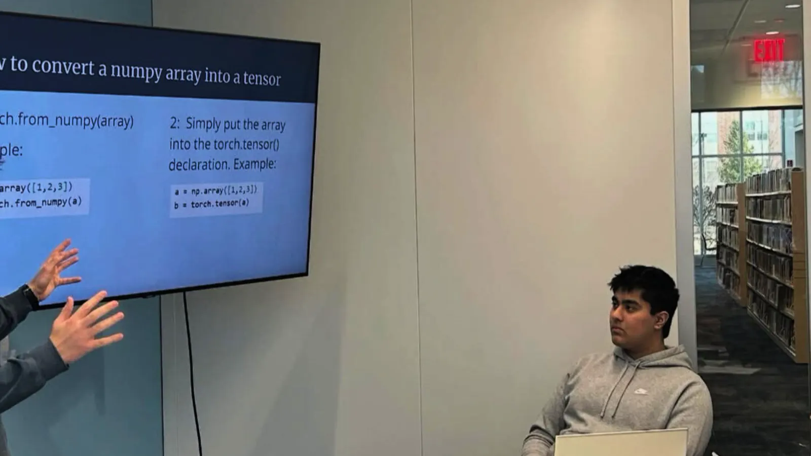 A presenter points to code examples on screen while a student listens, with library shelves behind.